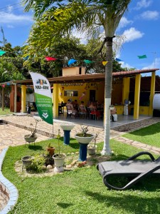 Área externa de uma chácara ensolarada, com gramado, piscina parcialmente visível, bandeirolas juninas, uma casa amarela ao fundo e banner do NU-RN próximo a pessoas reunidas à mesa.